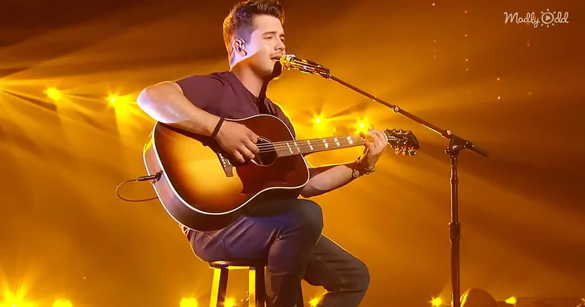A man sits on a stool, playing an acoustic guitar and singing into a microphone under warm stage lights.