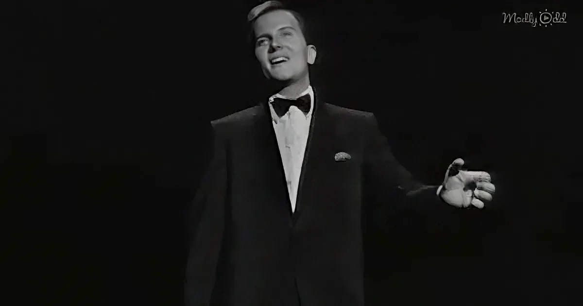 A man in a tuxedo and bow tie is singing, holding a microphone, against a dark background.