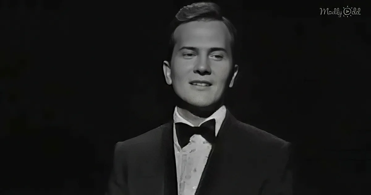 A man in a tuxedo and bow tie stands against a dark background, smiling.