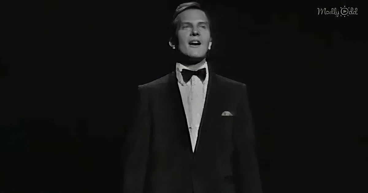 A man in a tuxedo sings against a dark background.