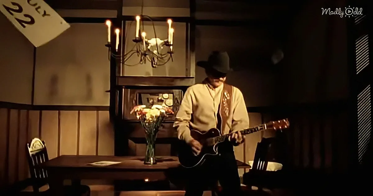 A man in a cowboy hat plays guitar in a dimly lit room with a chandelier and flowers.