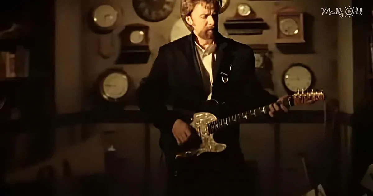 A man in a suit holds a guitar, standing in front of a wall with clocks.