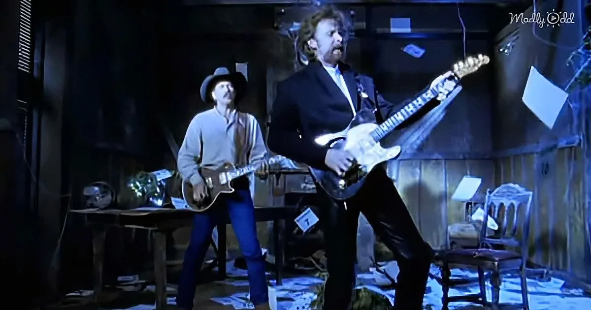 A man in a suit plays electric guitar passionately, another in a cowboy hat plays acoustic, both in a dimly lit...