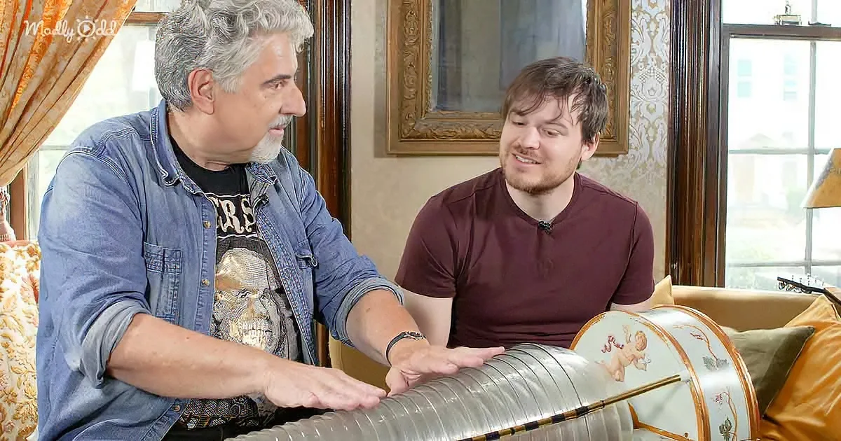 A man in a denim shirt interacts with a musical instrument while another man in a maroon shirt looks on.