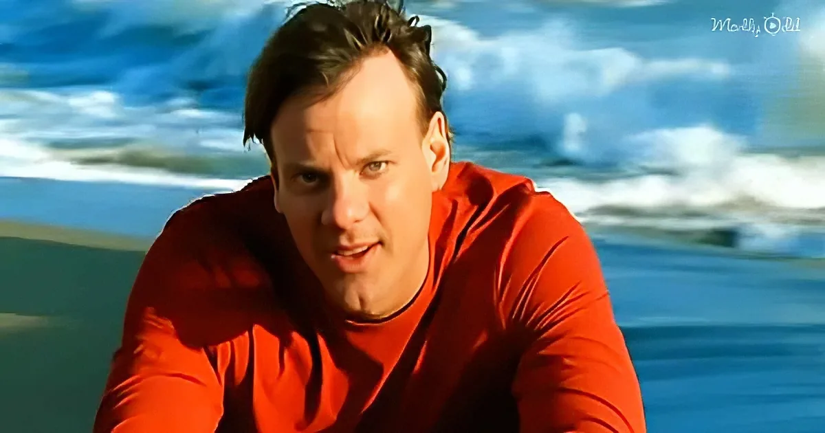 A man in a red shirt leans forward, looking intense, with a beach and ocean waves in the background.