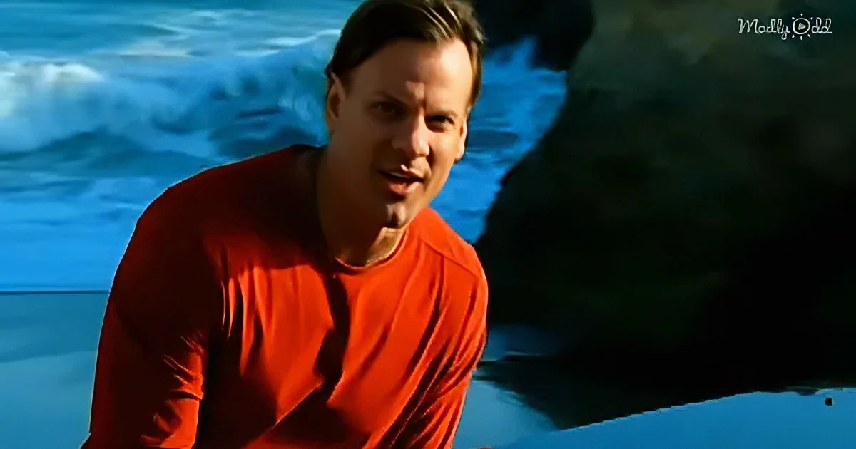 A man in a red shirt sits in front of a blue ocean backdrop, smiling.