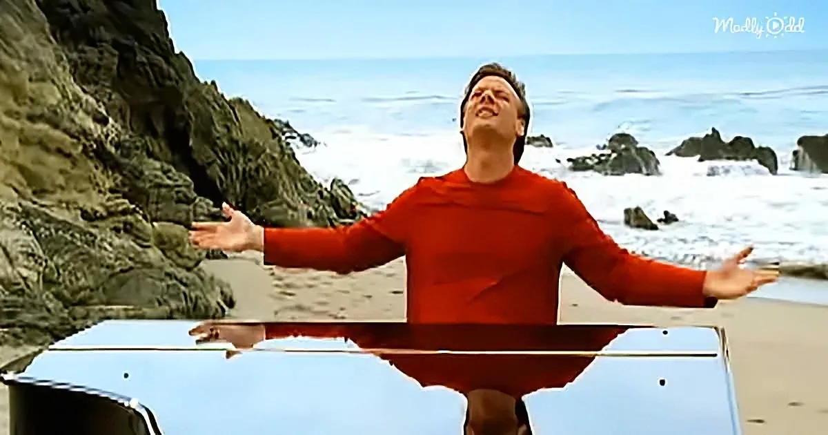 A man in a red shirt stands on a car hood with arms outstretched on a beach.