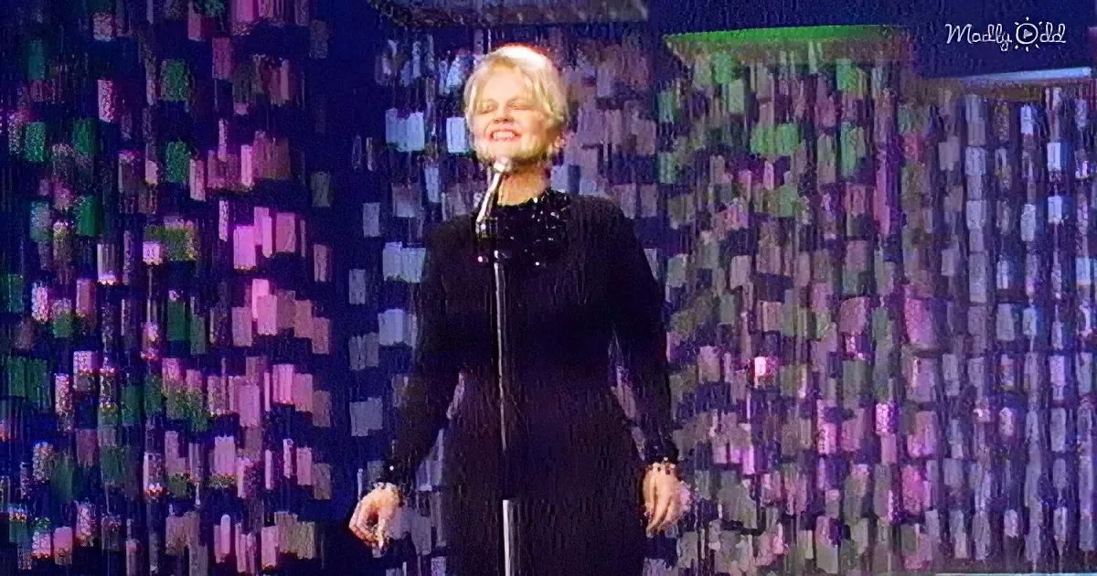 A person in a black outfit stands on stage with a microphone, surrounded by a colorful, patterned backdrop.