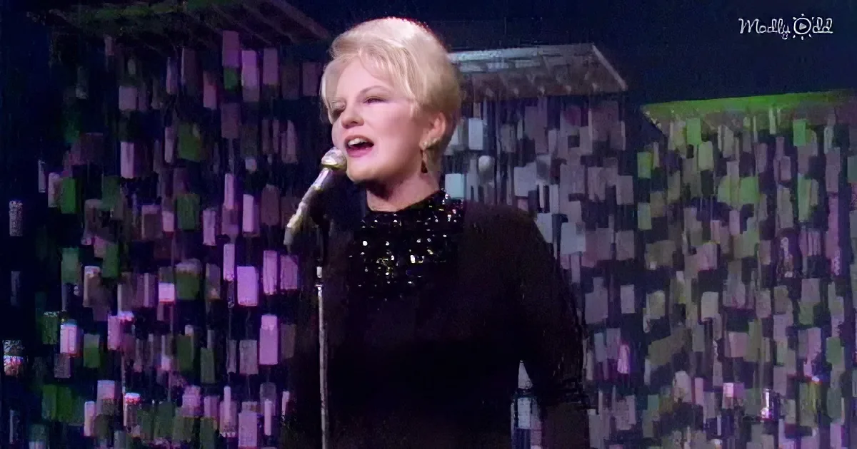 A woman in a black sequined dress sings into a microphone on stage with a colorful geometric backdrop.