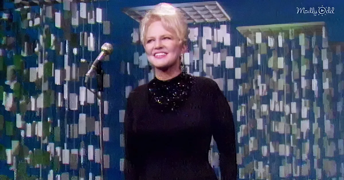 A woman in a black sequined dress smiles on stage with a microphone and patterned backdrop.