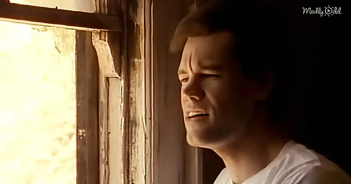 A man in a white shirt looks out a window with a distressed expression, sunlight streaming in.