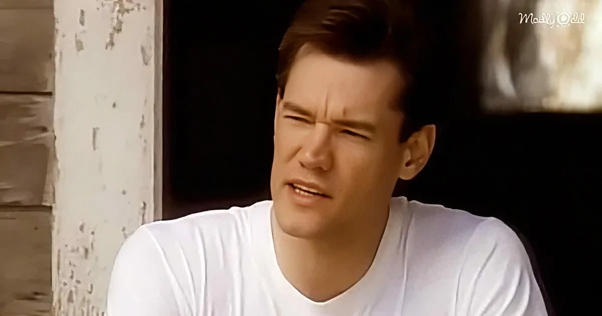 A man in a white t-shirt sits against a weathered wall, looking to the side with a slight smile.