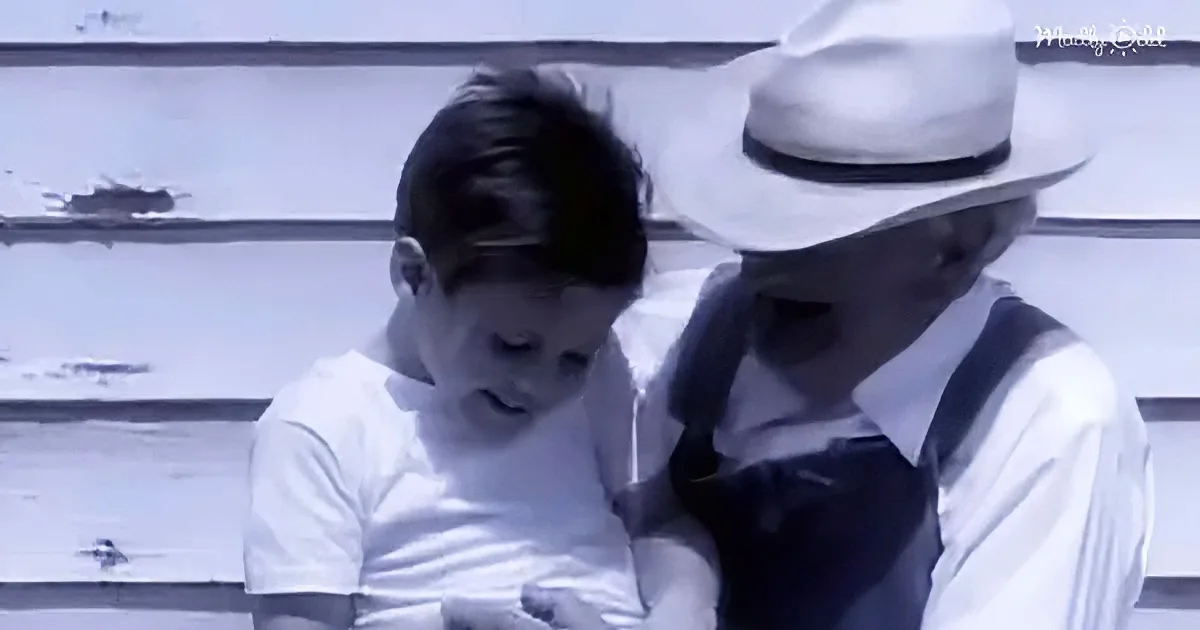 A young boy in a white shirt sits on a weathered bench, leaning against an older man in a white hat and suspenders.