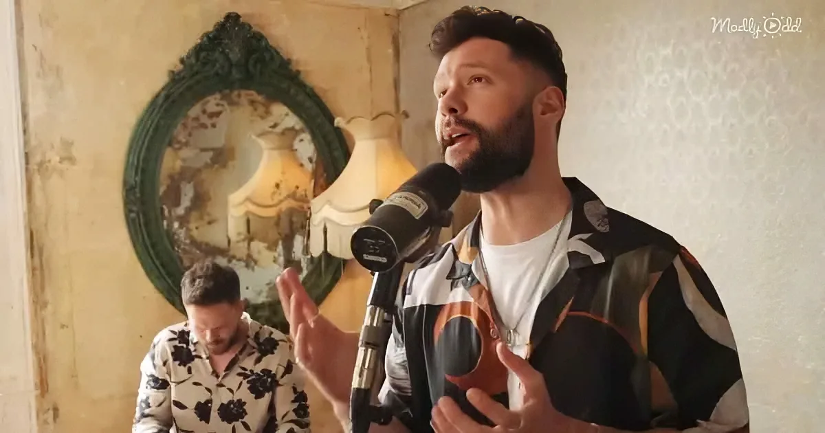A man with a beard speaks into a microphone, wearing a patterned shirt, with another person in floral attire in the...