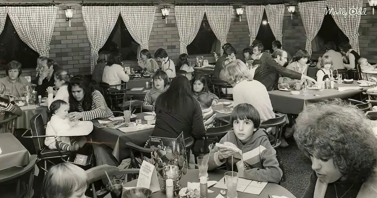 A boy sits at a table in a busy restaurant, surrounded by other diners, with a checkered curtain backdrop.