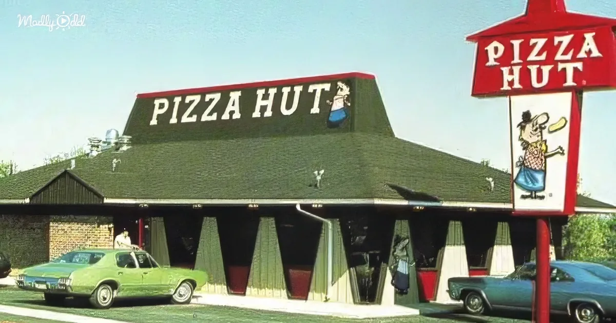 Pizza Hut restaurant with iconic red sign and cartoon character, vintage cars parked, green roof, and brick facade.