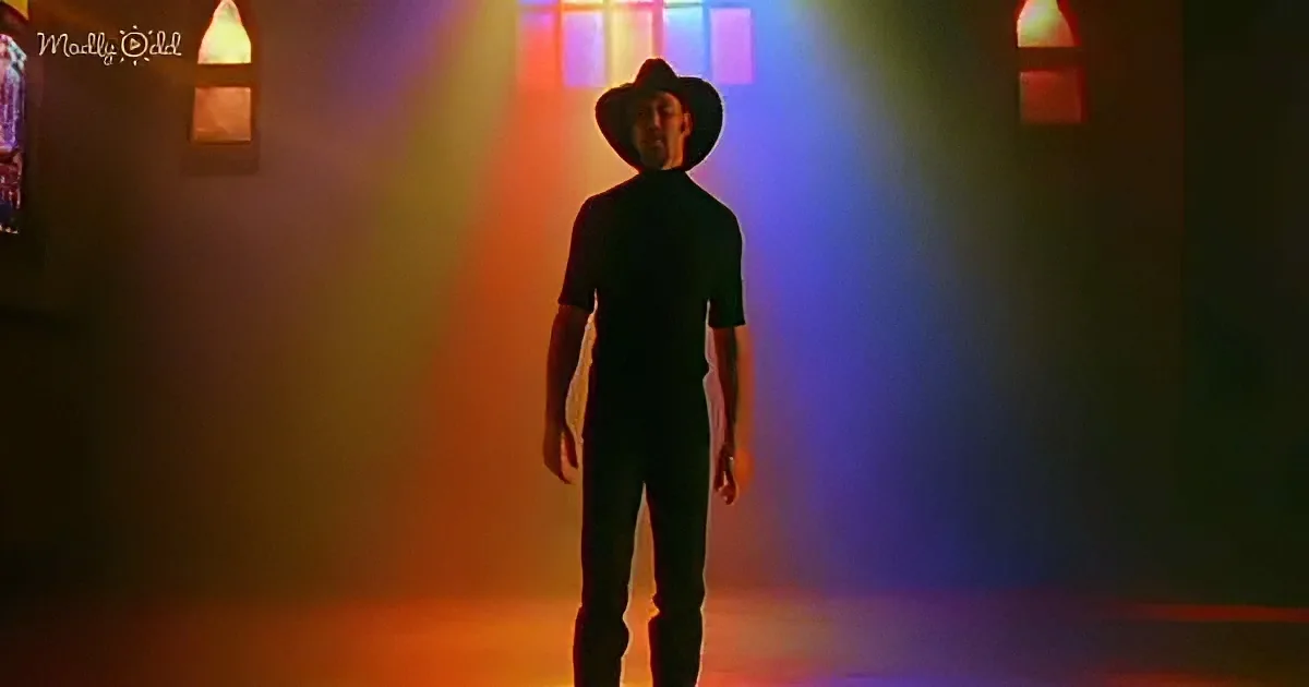 A man in a cowboy hat stands in a dimly lit room with colorful light beams behind him.