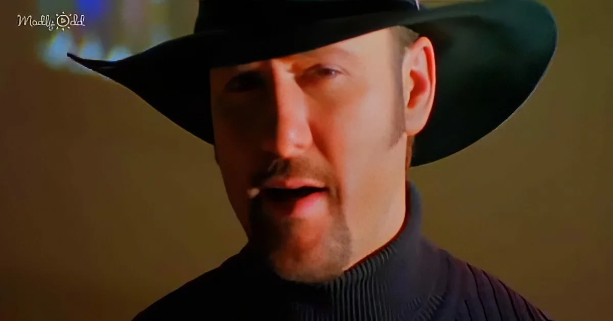A man wearing a black hat and turtleneck sweater is speaking with a blurred background.