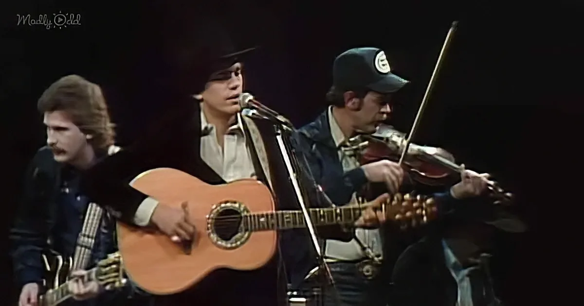 A man in a black suit plays an acoustic guitar on stage with a microphone and other musicians.