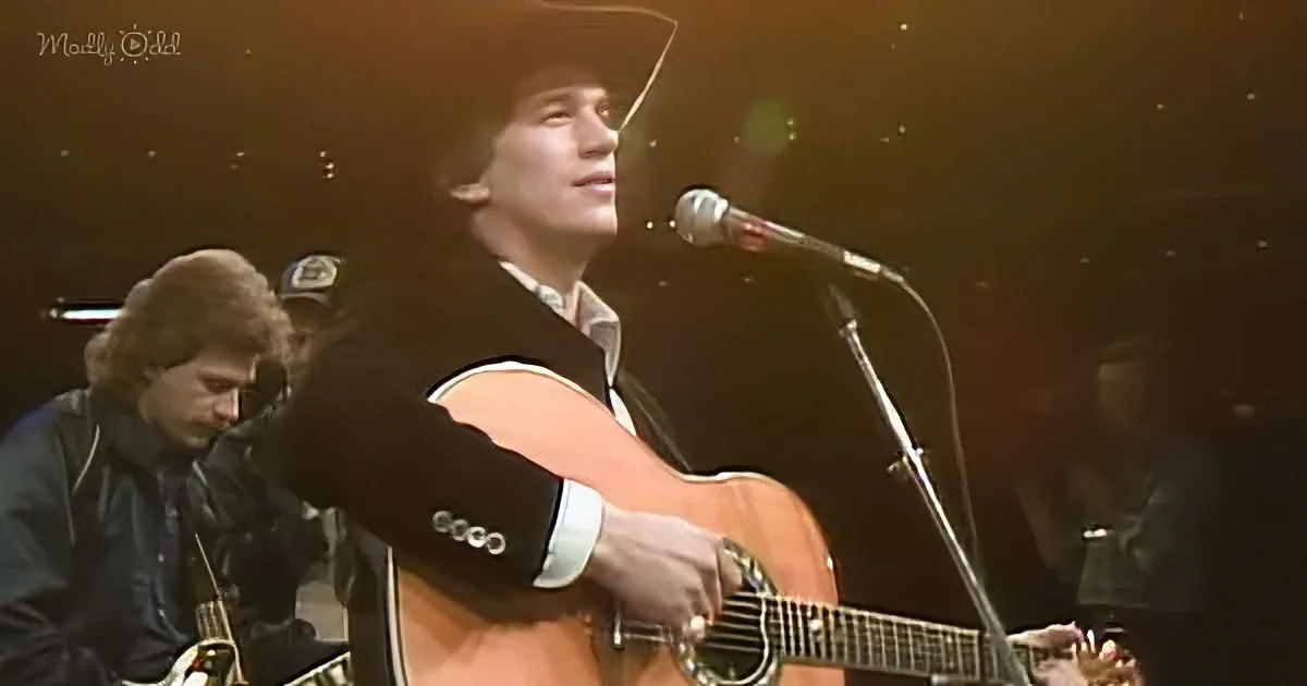 A man in a suit and cowboy hat plays an acoustic guitar on stage, singing into a microphone.