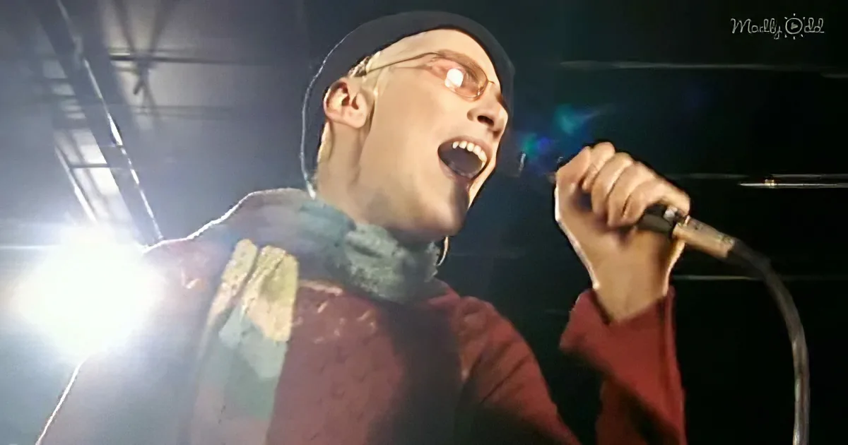 A person in a red sweater and scarf sings into a microphone under bright stage lights.