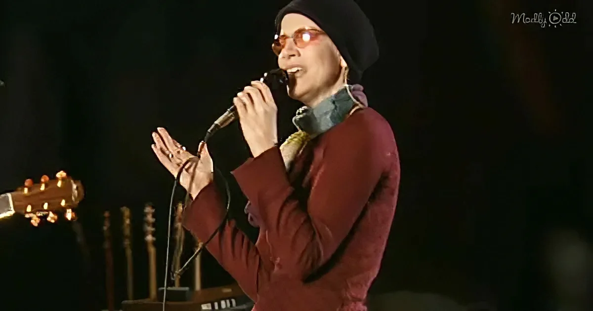A person wearing a beanie and red coat sings into a microphone, hands raised, with a guitar and amplifier in the background.