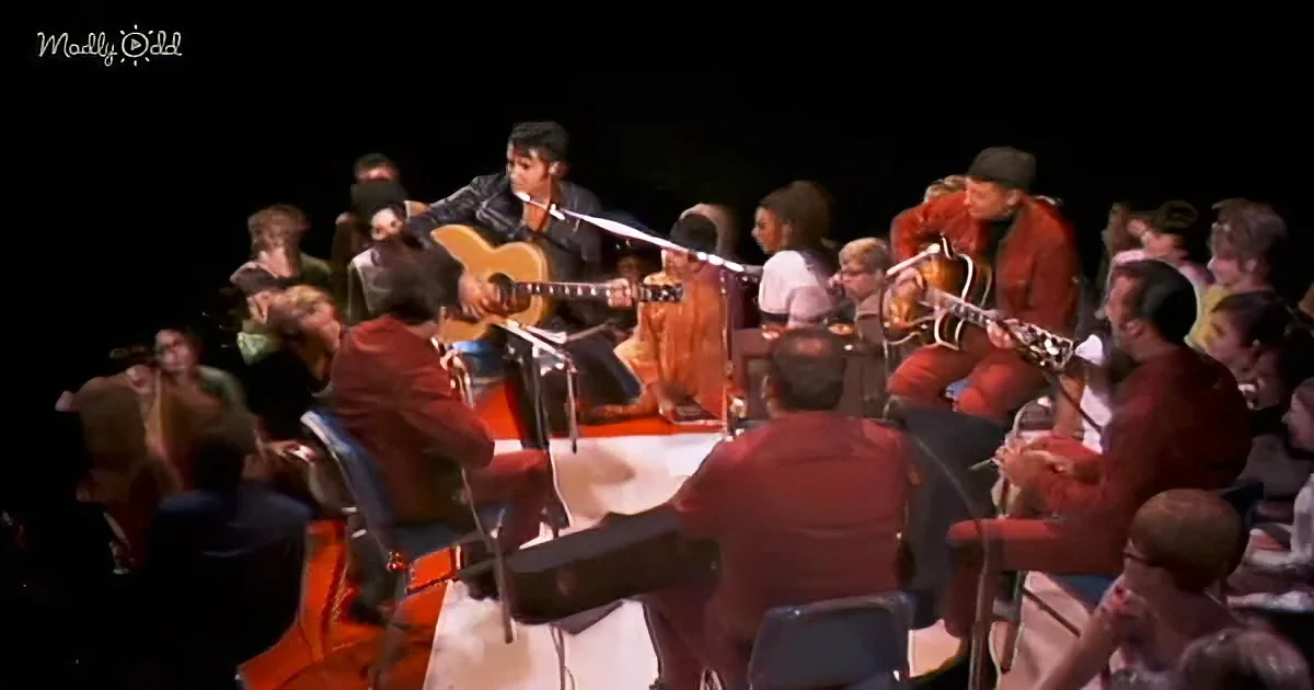 A man in a black jacket plays guitar on stage with an orchestra in red jackets.