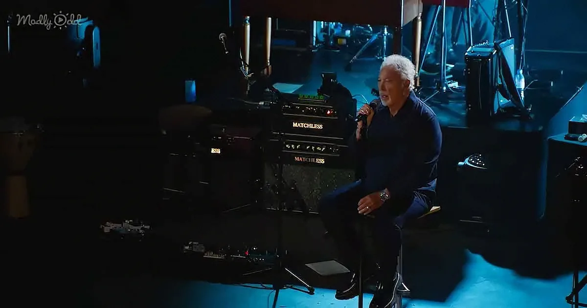 A man sits on stage, holding a microphone, surrounded by musical equipment and dim lighting.