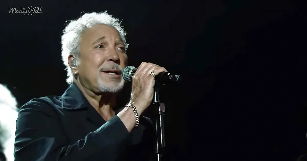 A man with white hair and a beard sings into a microphone on stage, wearing a black shirt and a bracelet.