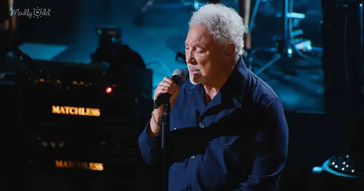 An older man with white hair and a mustache sings into a microphone on stage.