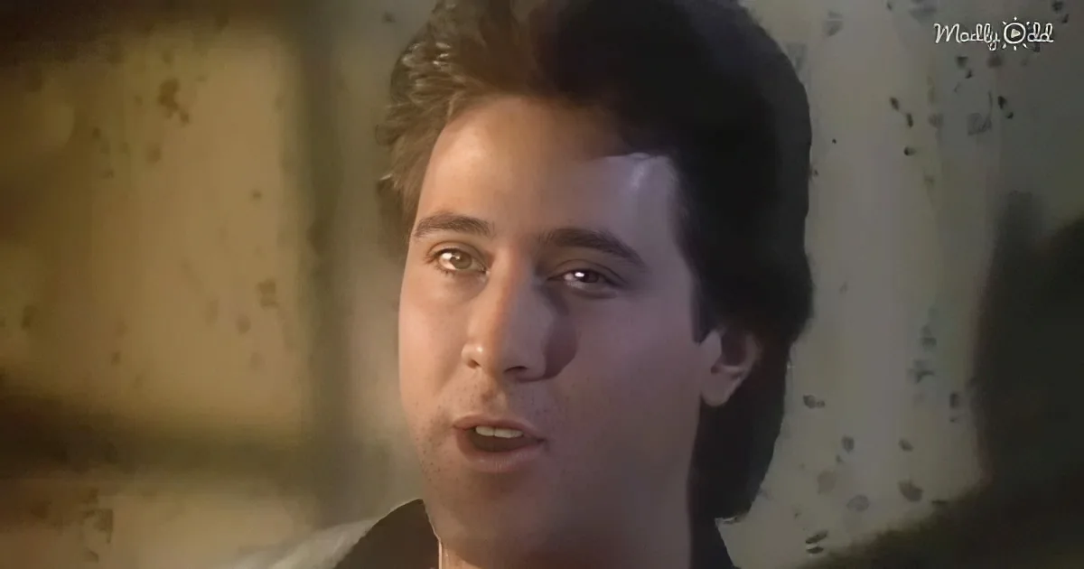 A man with dark hair and a light complexion is speaking, wearing a dark shirt, with a blurred background.