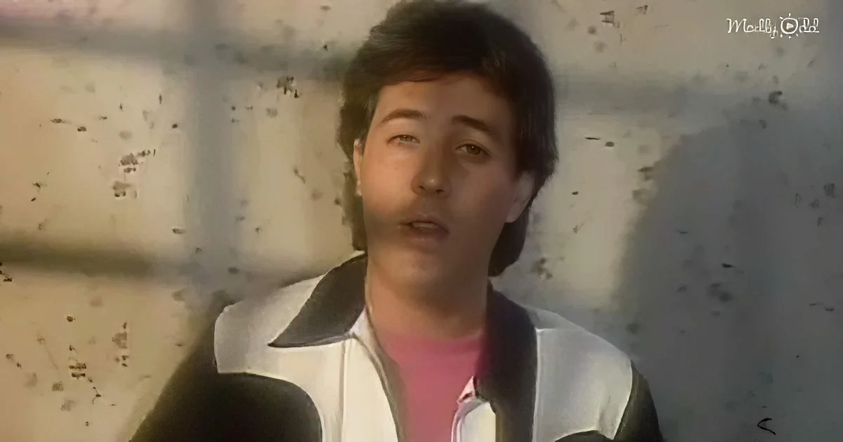 A person wearing a white jacket with a pink shirt underneath stands against a wall with dark spots.