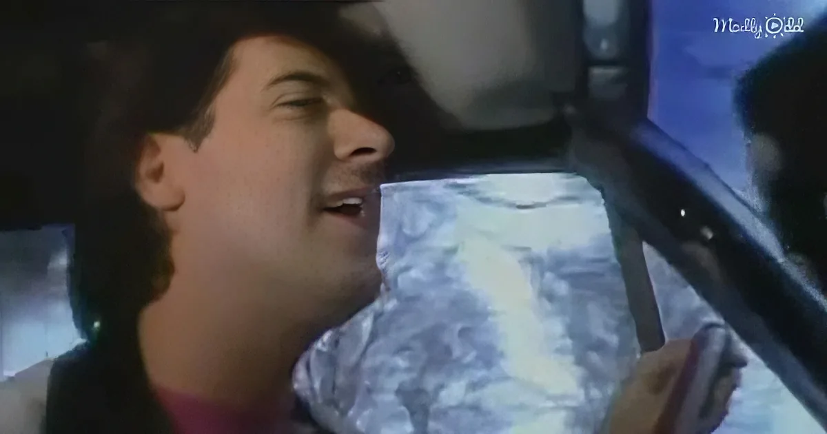 A person wrapped in foil, seemingly asleep, with a car interior and a hand holding a phone in the background.