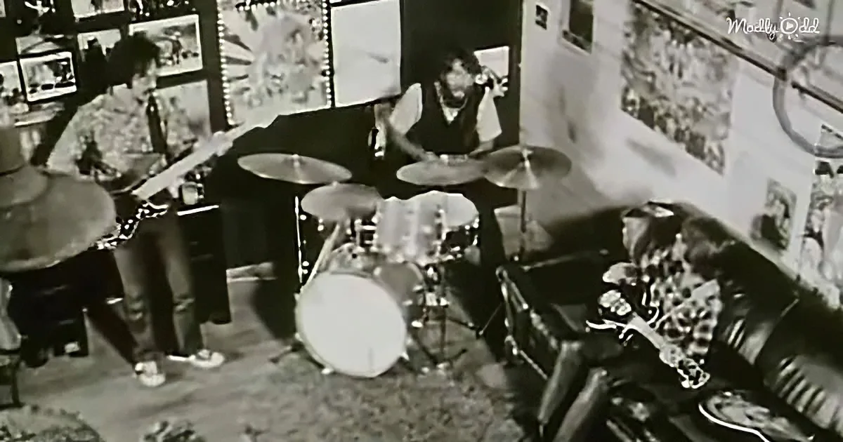 A band plays in a room with posters, a guitar player, a drummer, and a person on a chair.