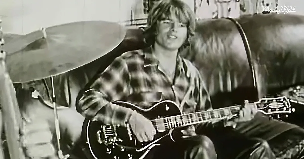 A person in a plaid shirt holds a guitar, smiling, with a drum set in the background.