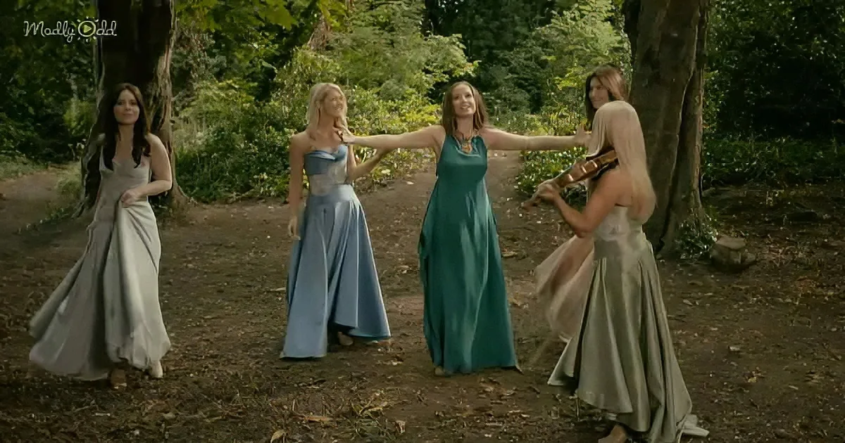 Four women in flowing dresses stand in a forest, one playing violin, another extending arms, others watching.