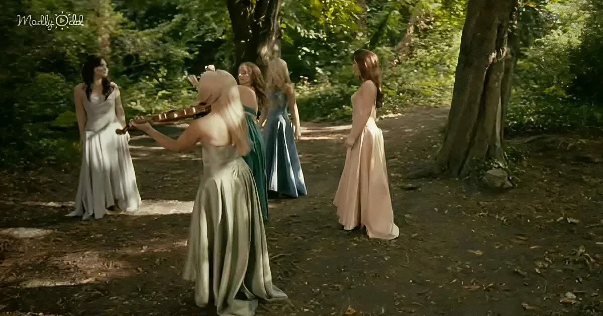 A woman in a white dress plays a violin in a forest, surrounded by four other women in elegant dresses.