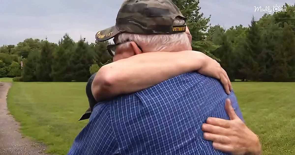 An elderly man in a plaid shirt and camouflage hat hugs another person in a park setting.