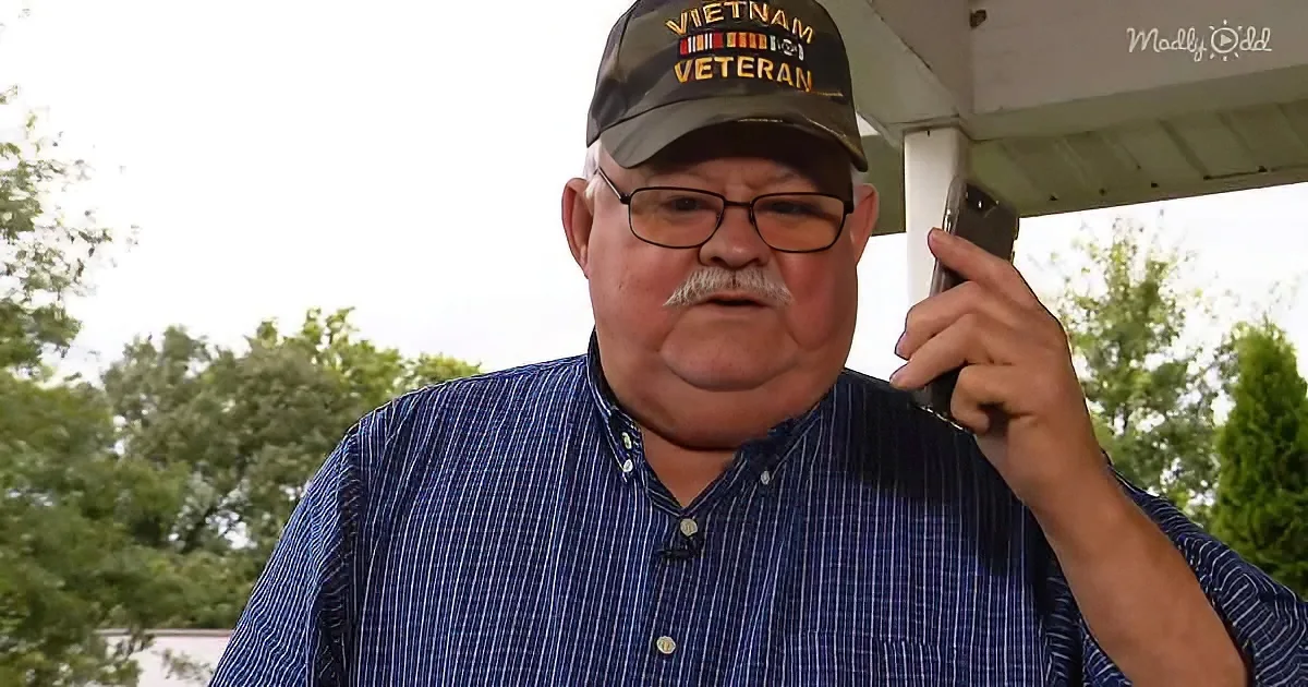 An older man wearing a "Vietnam Veteran" cap and glasses holds a phone to his ear, standing outdoors.