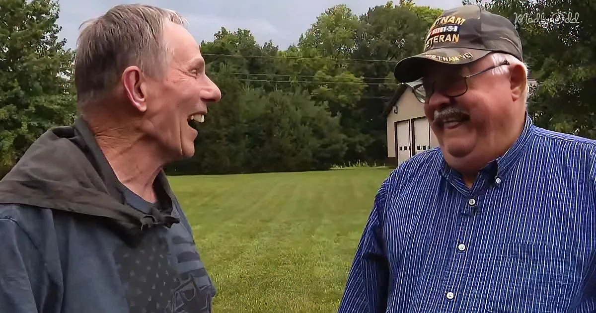 Two men, one in a "Vietnam Veteran" hat, share a laugh in a grassy field with trees and a building in the background.