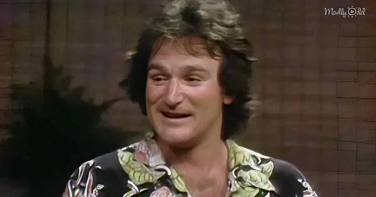 A man with curly hair, wearing a floral shirt, smiles warmly in front of a brick wall.