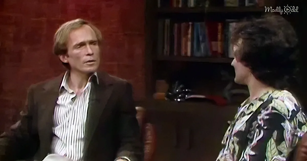 A man in a suit and striped shirt sits, facing another person in a floral shirt, in a room with a bookshelf.