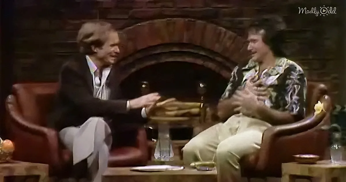 Two men in a cozy setting, one in a suit, the other in a patterned shirt, shaking hands across a table.
