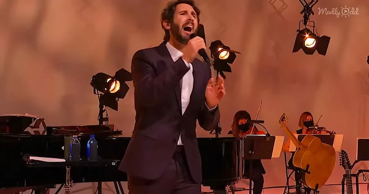 A man in a suit sings passionately into a microphone on stage, with a band and spotlights behind him.