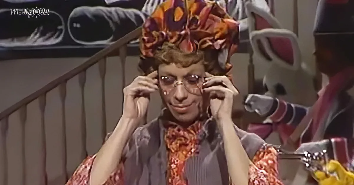 A person in a colorful outfit and elaborate headpiece adjusts their glasses, standing indoors near stairs.