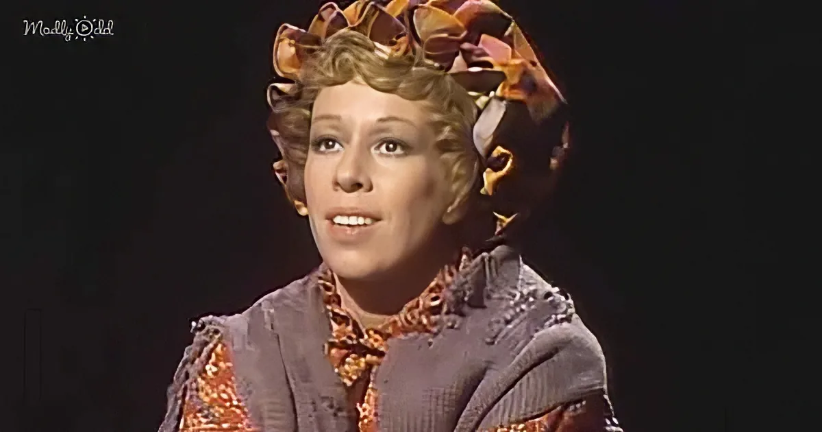 A woman in a vintage-style outfit with a floral headpiece and patterned blouse smiles against a dark background.