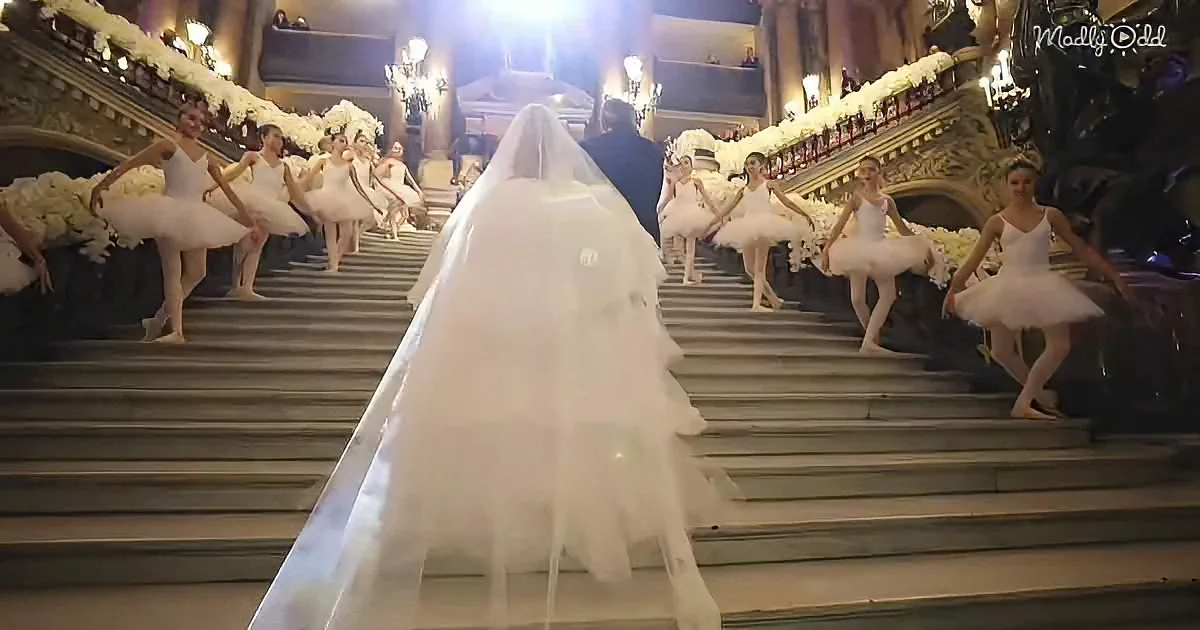 A bride descends grand stairs flanked by ballerinas in white tutus, under ornate chandeliers.