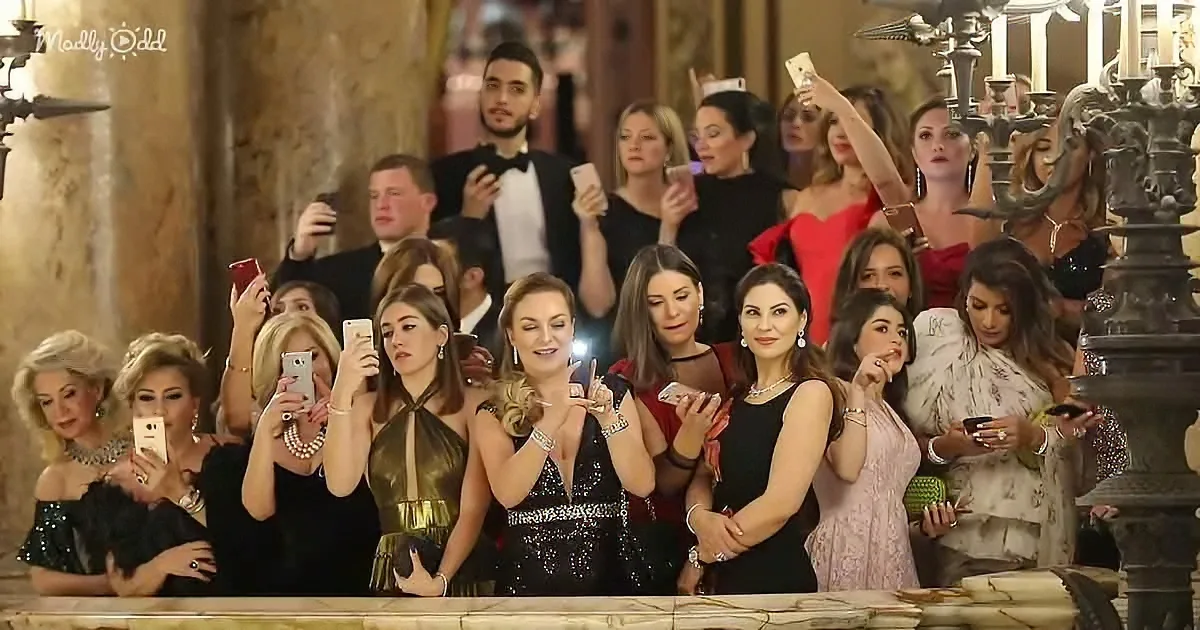 A group of elegantly dressed women are taking photos with their phones in a grand, ornate setting.