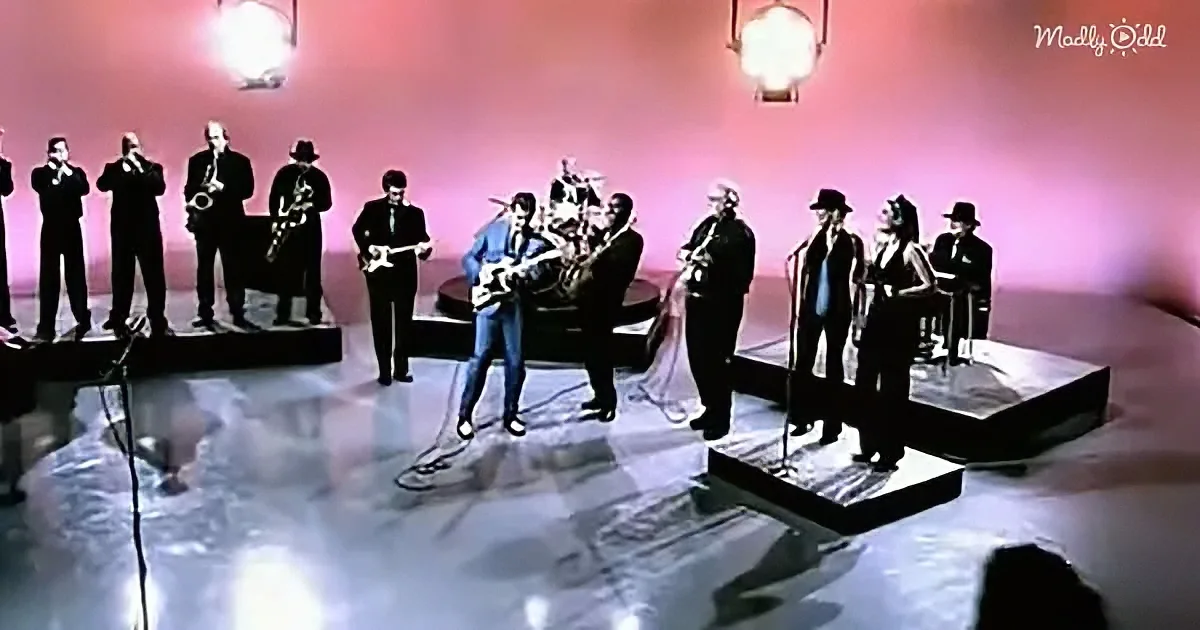 A man in a blue suit plays guitar on stage surrounded by musicians.