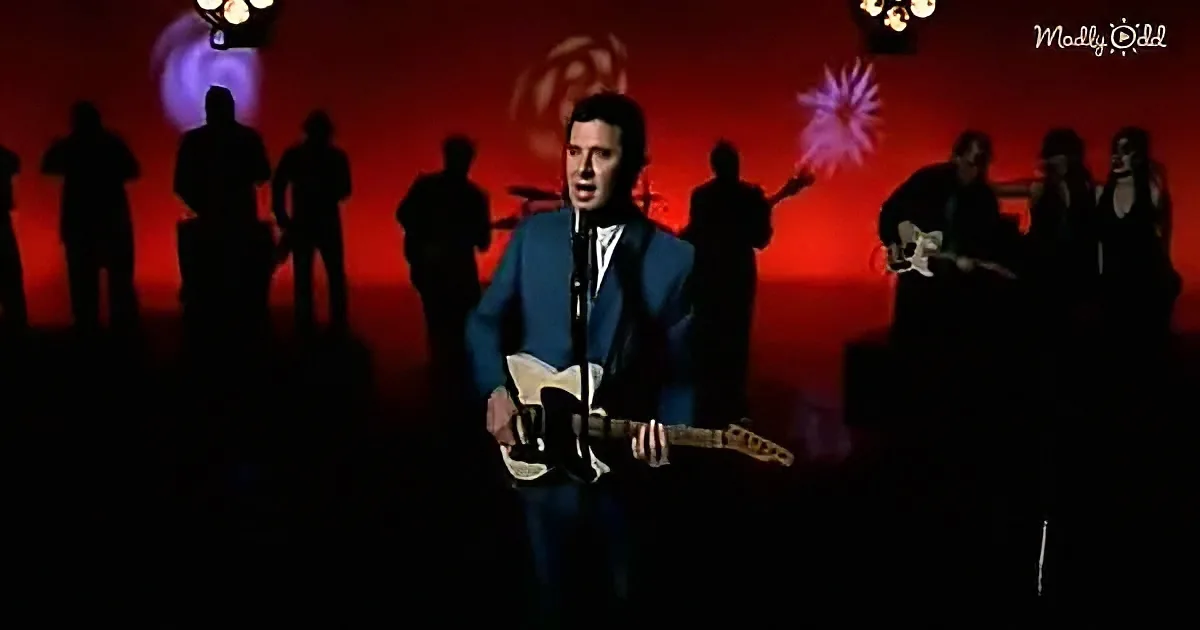 A man in a blue suit sings into a microphone while holding a guitar, with a band and colorful lights in the background.